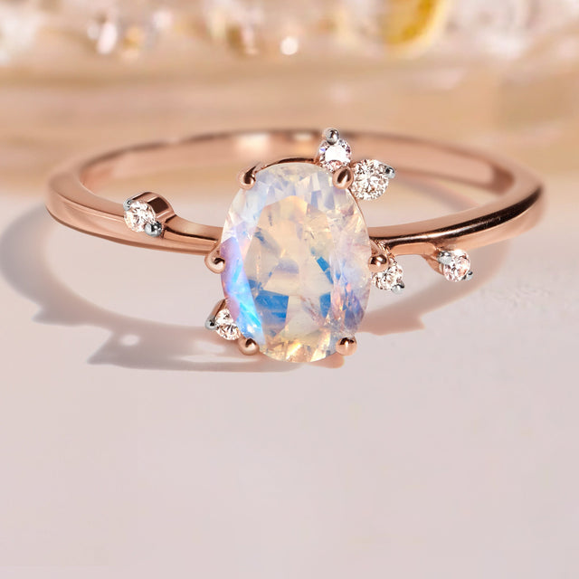 A rose gold moonstone ring rests on a pale tabletop with blurred glass behind it.