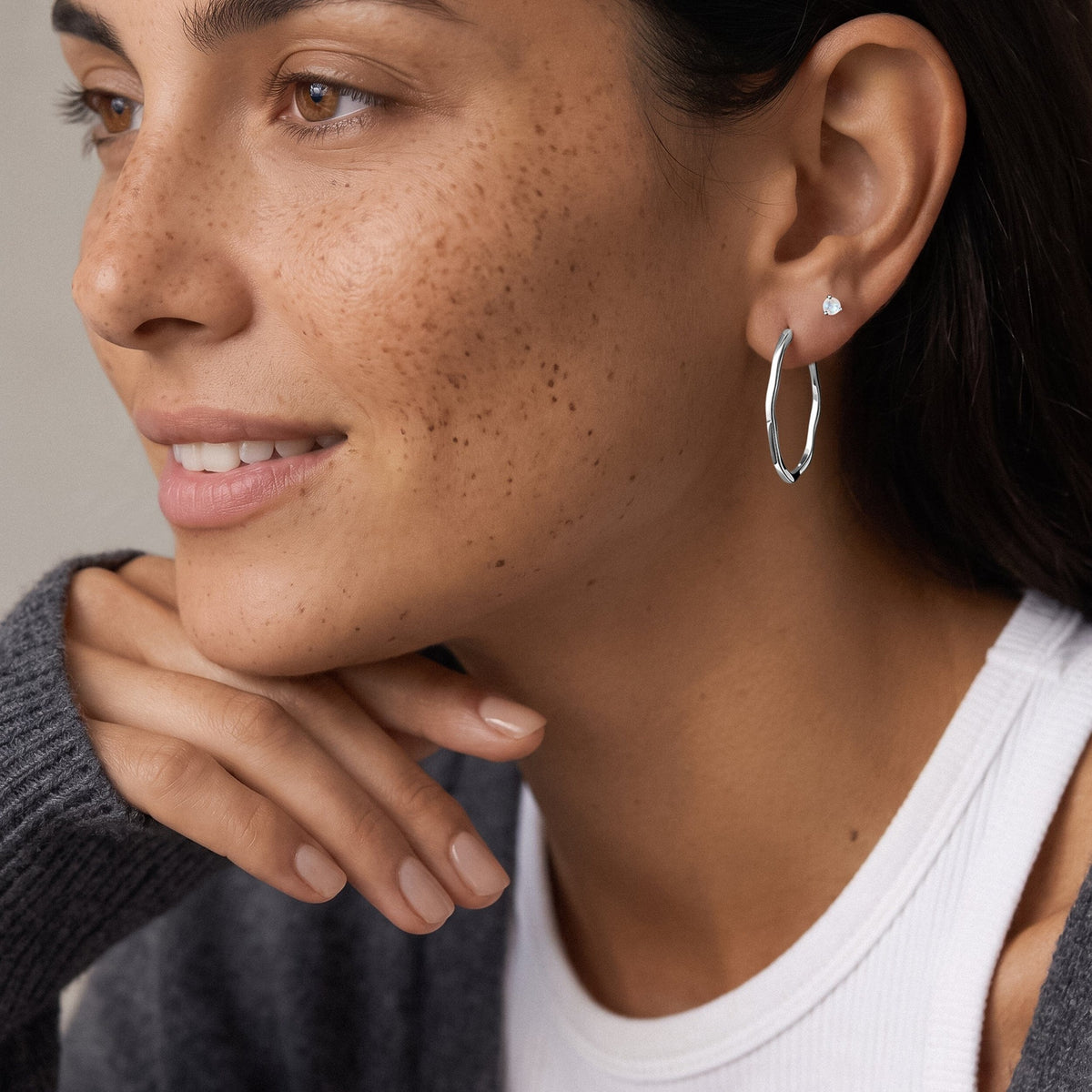 Smiling freckled person wears an elongated wavy hoop earring with a small round stud above it.