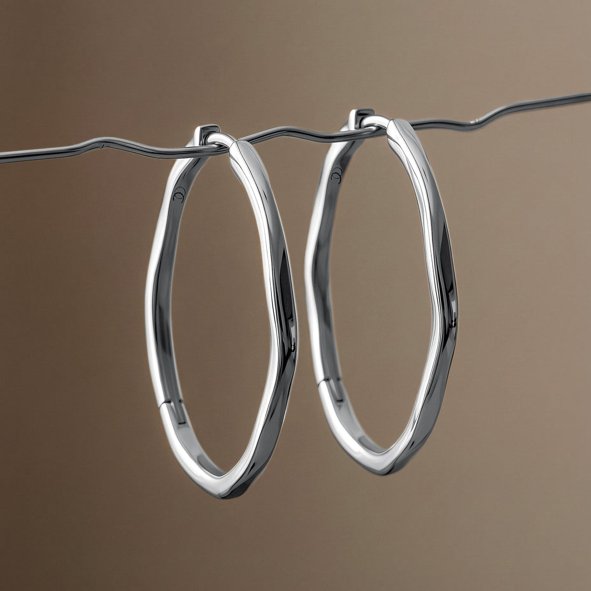Pair of elongated wavy hoops hang from a thin wavy line against a softly blurred warm brown background.