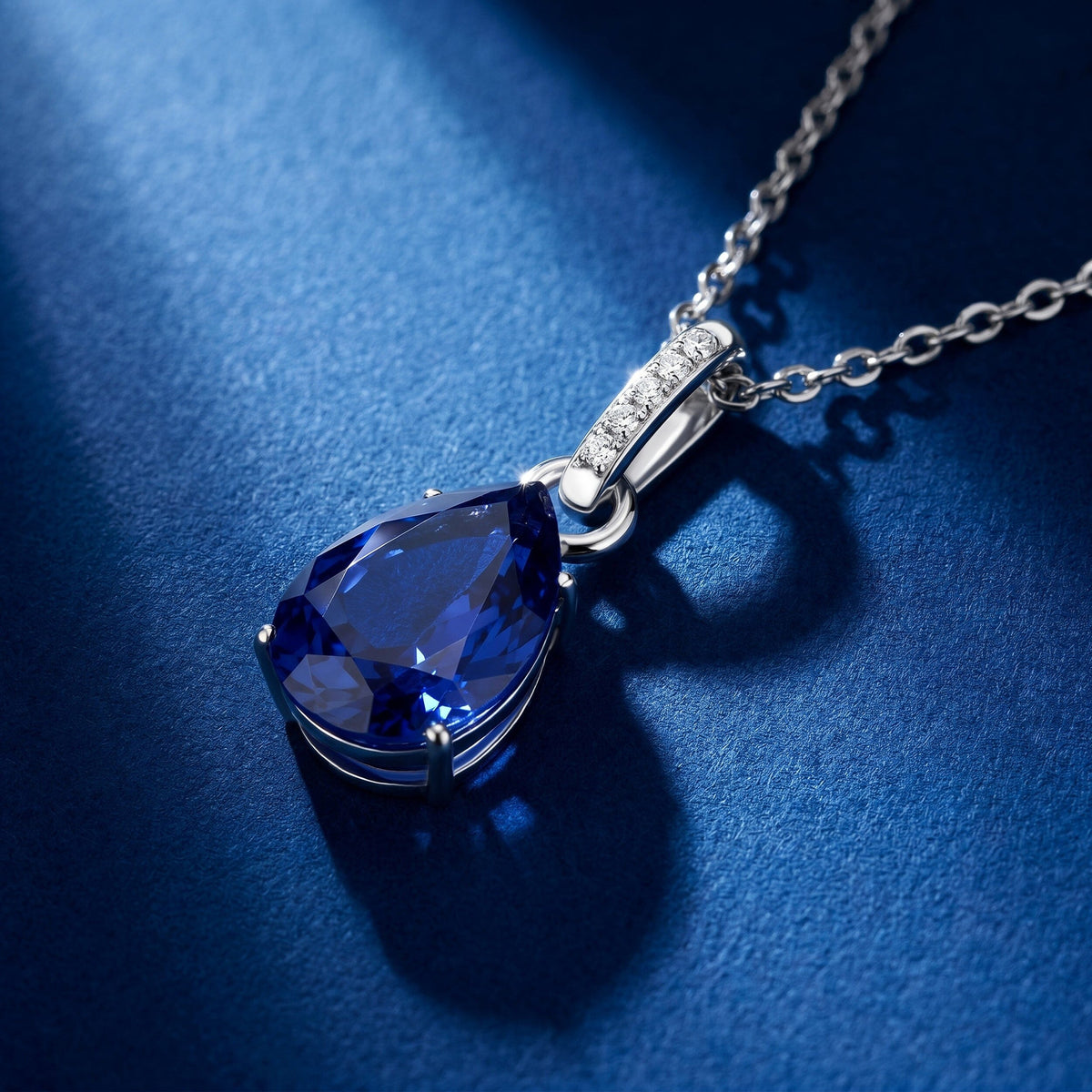 A silver necklace rests on a blue textured tabletop with the sapphire and curved bail seen at an angle.