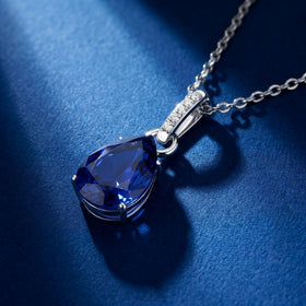A silver necklace rests on a blue textured tabletop with the sapphire and curved bail seen at an angle.