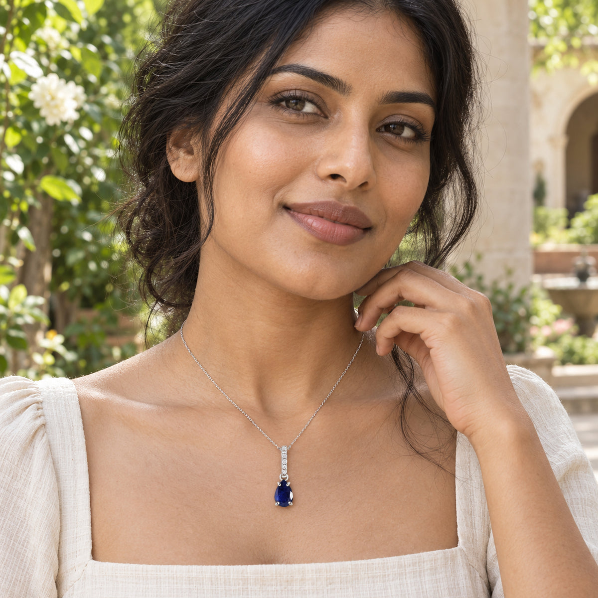 A woman in a cream square-neck top wears the silver blue sapphire pendant in a garden courtyard.