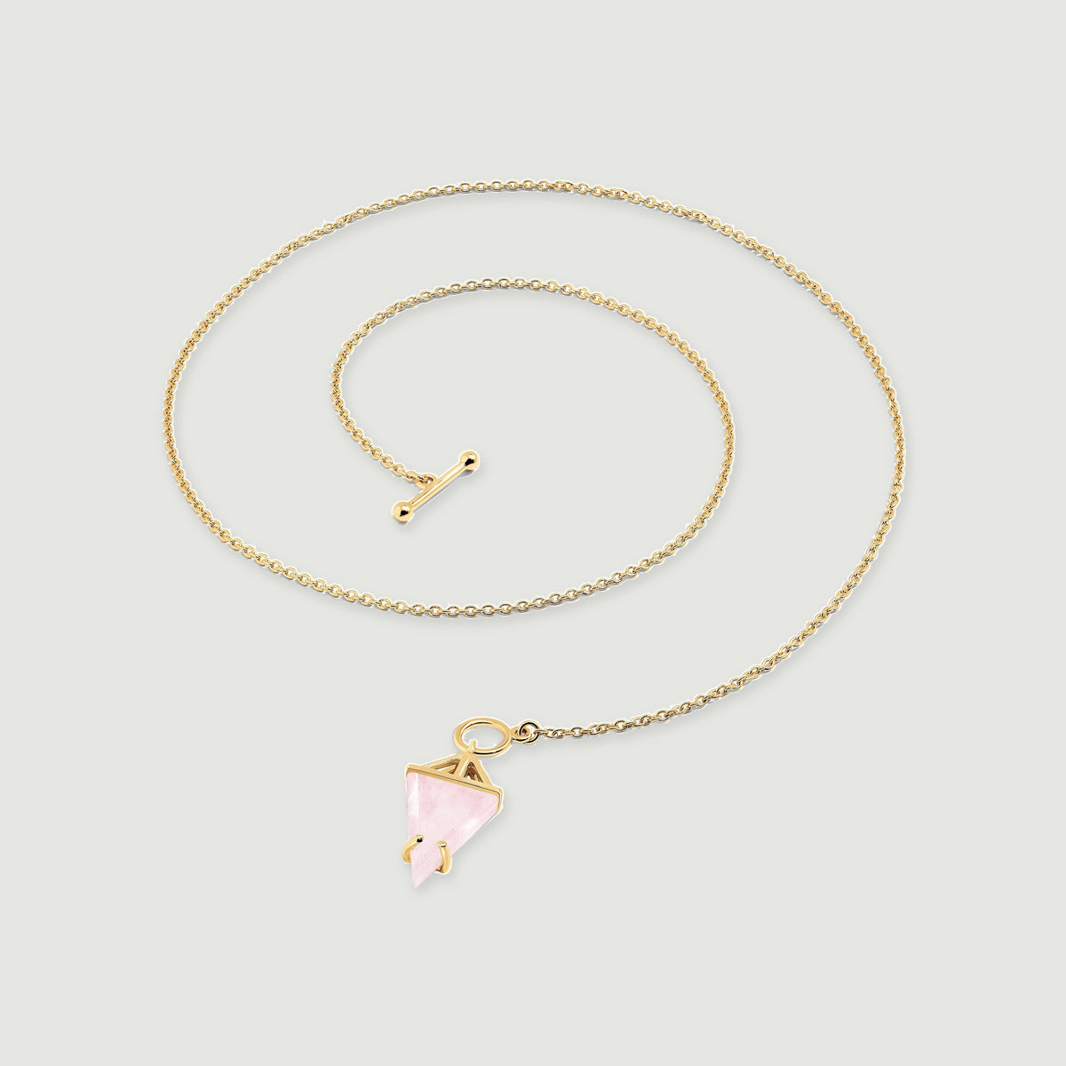 A yellow gold necklace is laid in a curved coil with a pink triangular stone and a separate toggle bar.