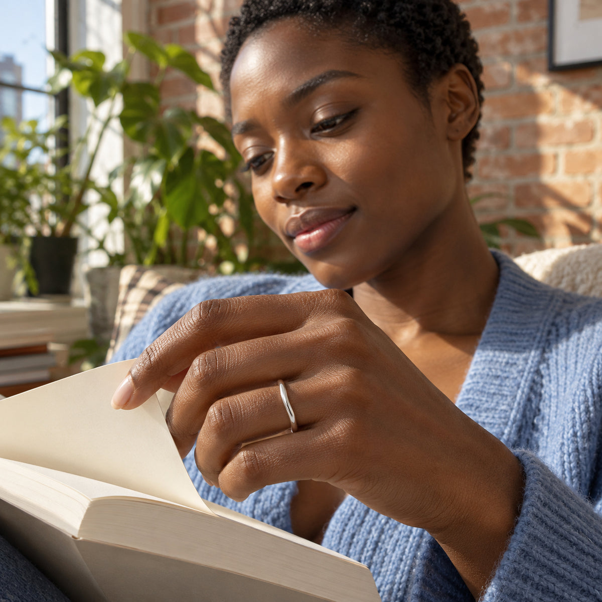 A polished silver band is worn on a hand turning a page beside a sunlit window.