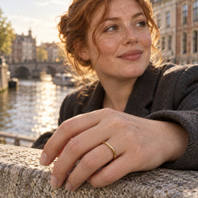 A yellow gold band sits on a hand resting on a stone ledge beside a canal.