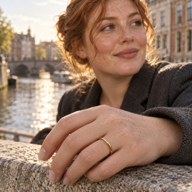 A yellow gold band sits on a hand resting on a stone ledge beside a canal.