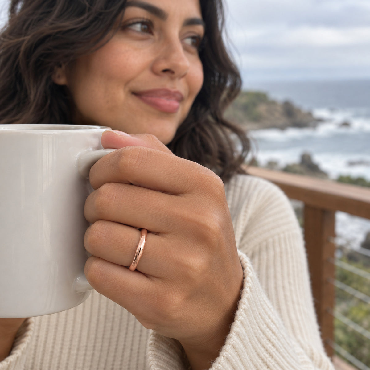 A rose gold band is shown on a hand holding a white mug with a coastal view behind it.