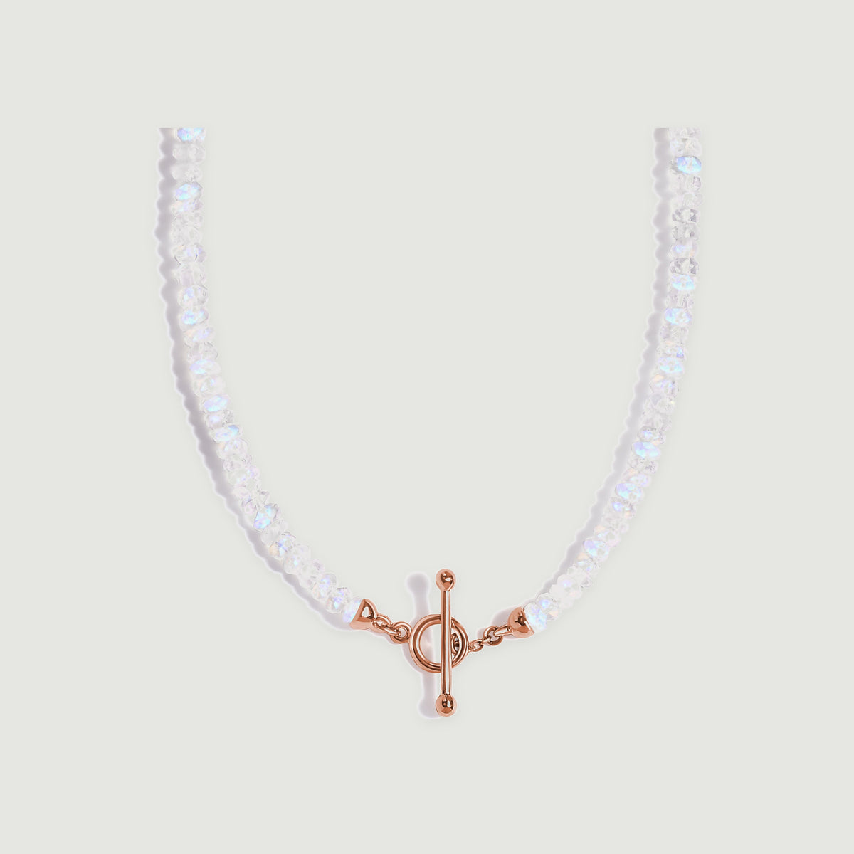 A curved strand of faceted moonstone beads meets a rose gold bar and circular clasp on a light grey background.