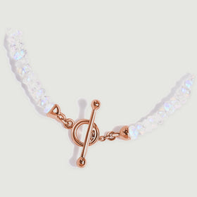 The lower curve of the moonstone necklace rests diagonally with its rose gold clasp against a grey surface.
