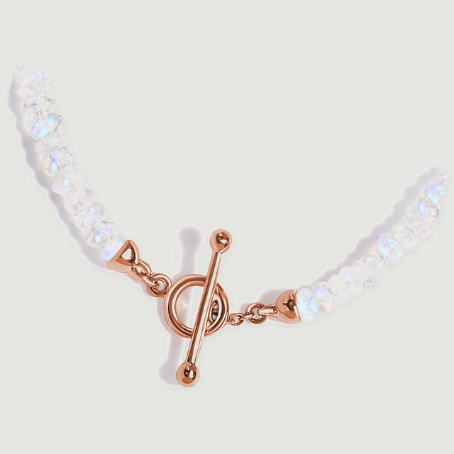 The lower curve of the moonstone necklace rests diagonally with its rose gold clasp against a grey surface.