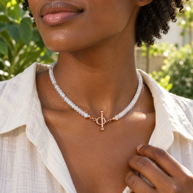 A woman in a cream linen shirt wears the moonstone bead necklace with a rose gold front clasp in a leafy courtyard.