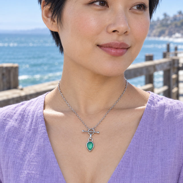A woman in a lavender top wears a silver-toned necklace with a green pear pendant beside a seaside pier.