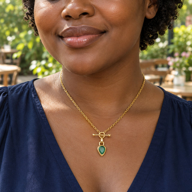 A woman with short curls wears the gold-toned green stone necklace above a navy blouse at an outdoor cafe.