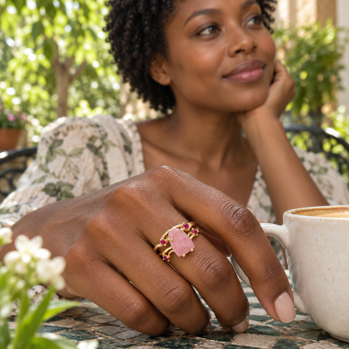 A woman wears a yellow gold vermeil rose quartz and ruby ring beside a coffee cup in a leafy courtyard.