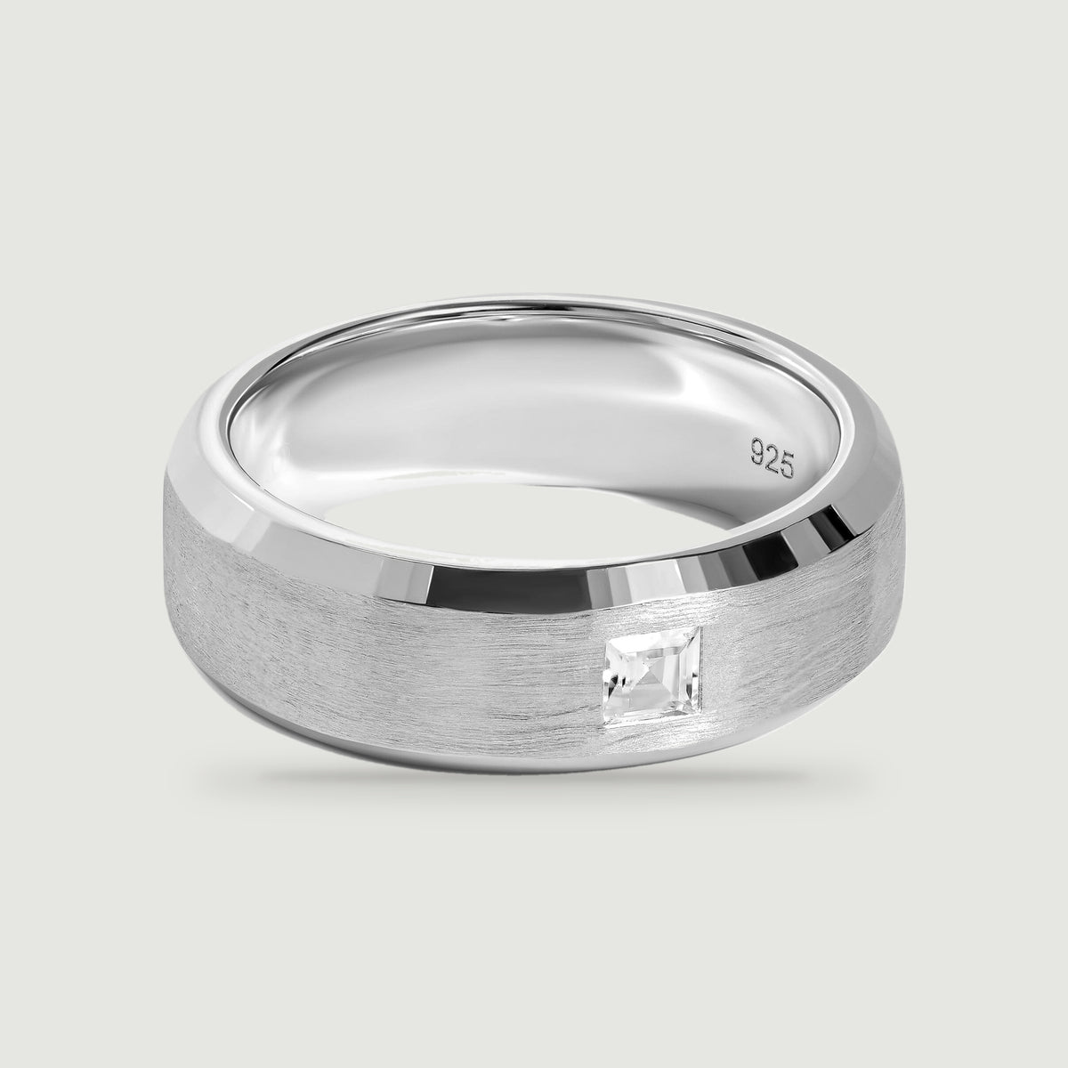 A silver ring with a brushed band and a single square clear stone rests on a light gray surface.