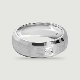 A silver ring with a brushed band and a single square clear stone rests on a light gray surface.