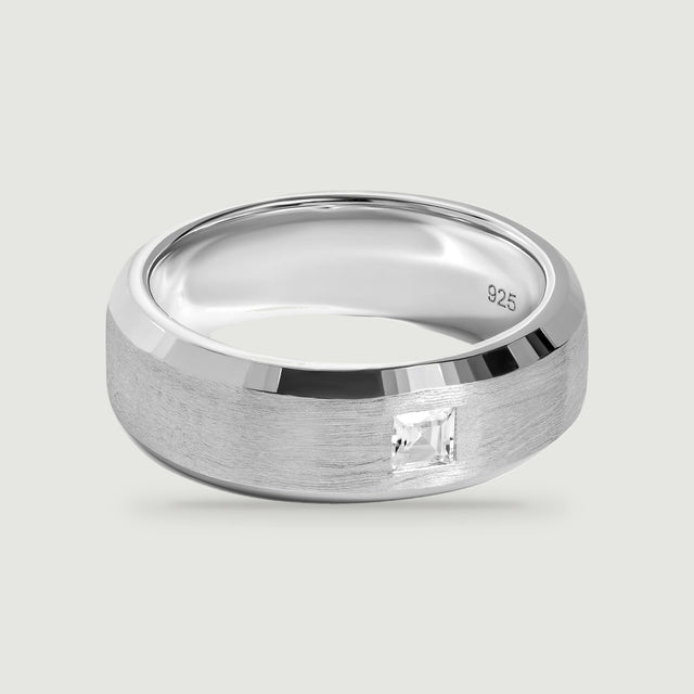 A silver ring with a brushed band and a single square clear stone rests on a light gray surface.