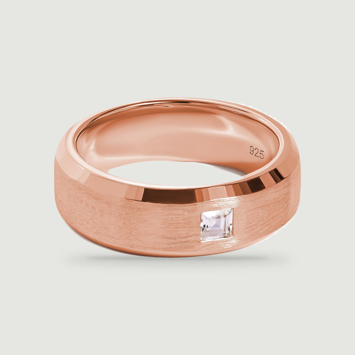 A rose gold ring with a wide textured band and a square clear stone is shown from above.