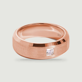 A rose gold ring with a wide textured band and a square clear stone is shown from above.
