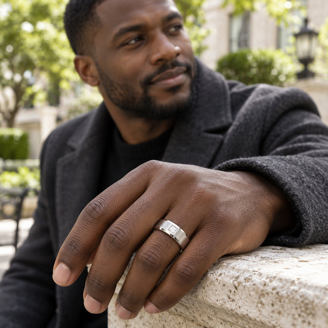 A silver ring with a square clear stone is worn on a hand resting on a pale stone railing.