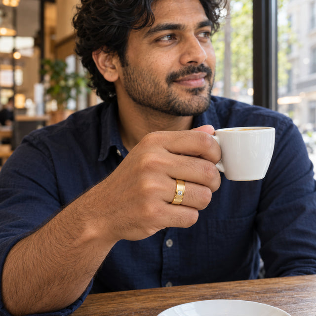 A yellow gold ring with a brushed band and square clear stone is worn while holding a white ceramic cup.