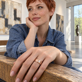 A rose gold ring with a square clear stone is shown on a freckled hand resting on a wooden bench.