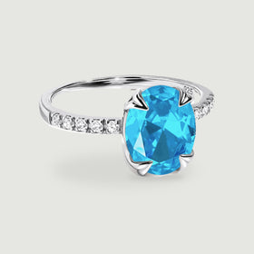 A sterling silver ring with an oval blue gemstone and clear stones along the band rests on a light grey surface.