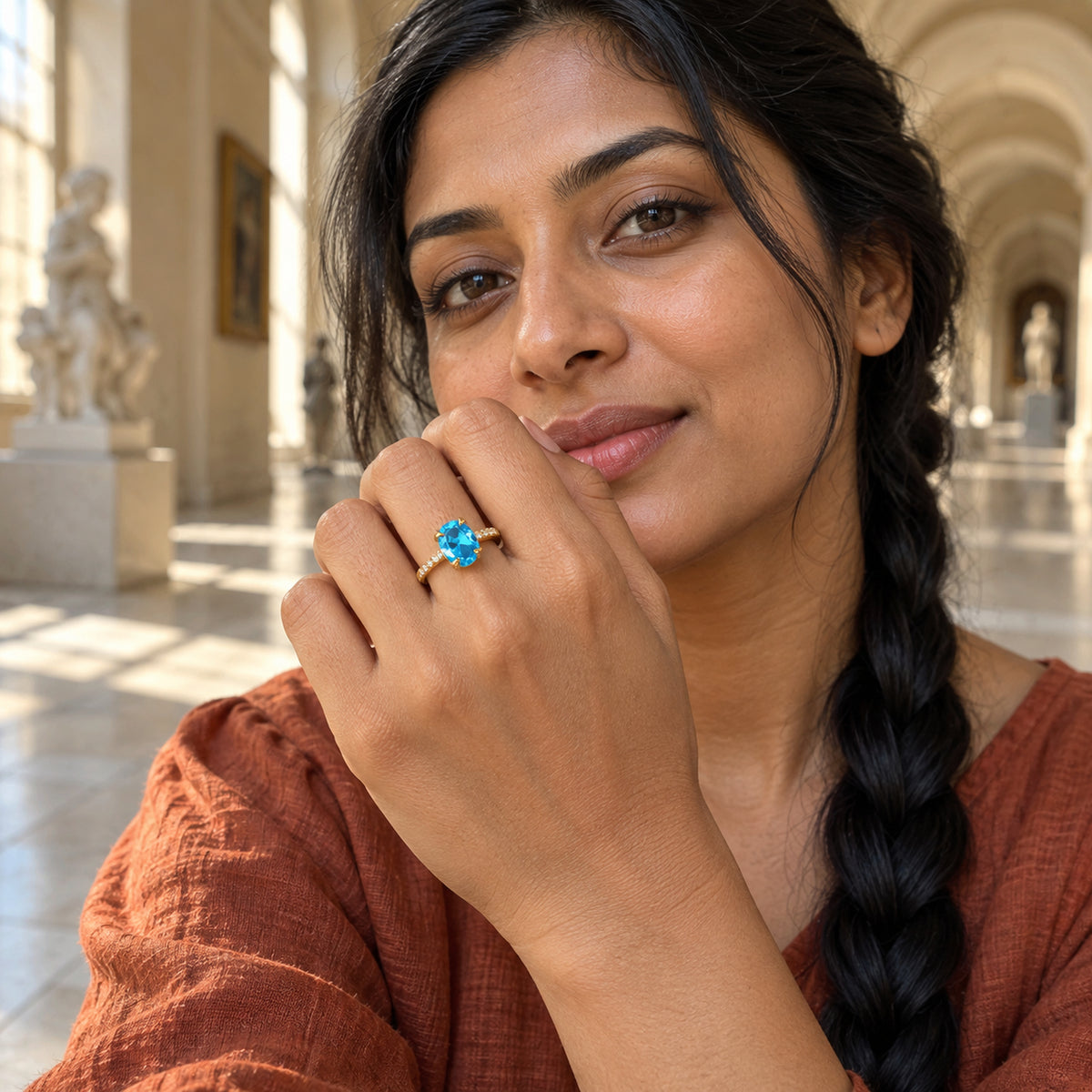 A woman with a long dark braid shows a yellow gold ring with a blue oval gemstone inside a sunlit arched gallery.