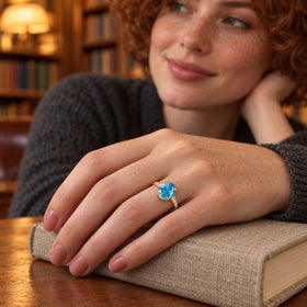 A freckled hand wearing a yellow gold ring with a blue gemstone rests on a linen-covered book beside wooden shelves.
