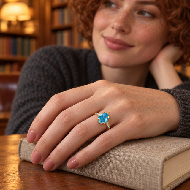 A freckled hand wearing a yellow gold ring with a blue gemstone rests on a linen-covered book beside wooden shelves.