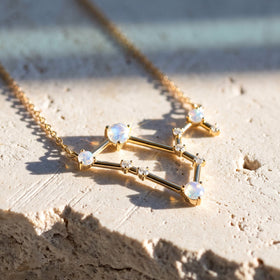 A yellow gold constellation necklace lies diagonally on a pale textured stone slab.