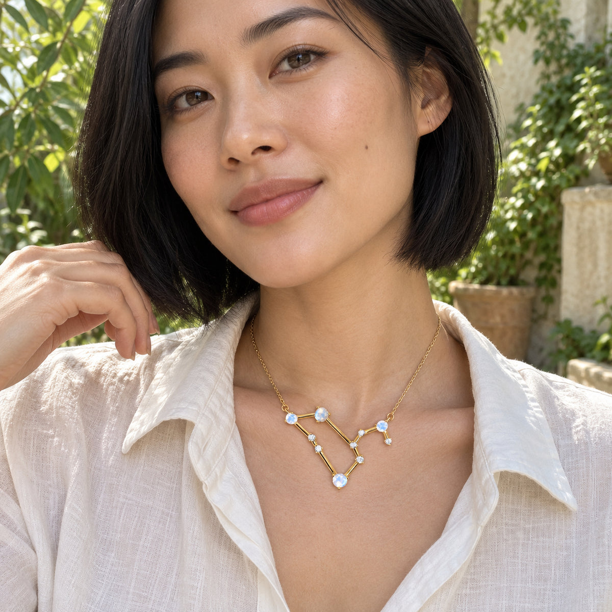 A woman in a white linen shirt wears a yellow gold moonstone necklace in a leafy courtyard.