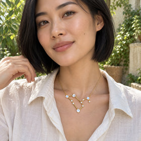 A woman in a white linen shirt wears a yellow gold moonstone necklace in a leafy courtyard.