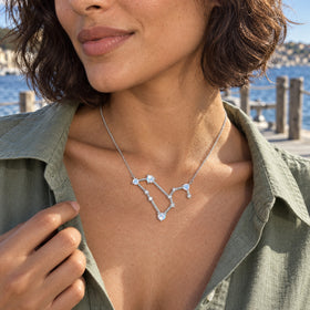 A silver moonstone constellation necklace rests below the collarbones of a woman in a green shirt beside the water.