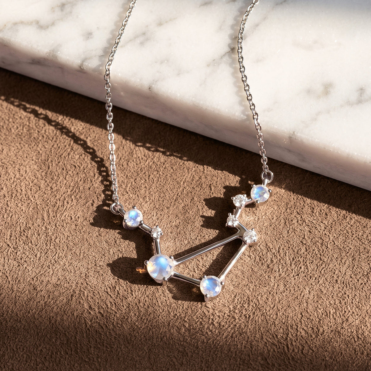 A silver constellation necklace lies across a textured brown surface beside a pale marble slab.