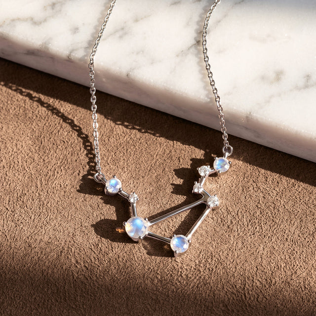 A silver constellation necklace lies across a textured brown surface beside a pale marble slab.