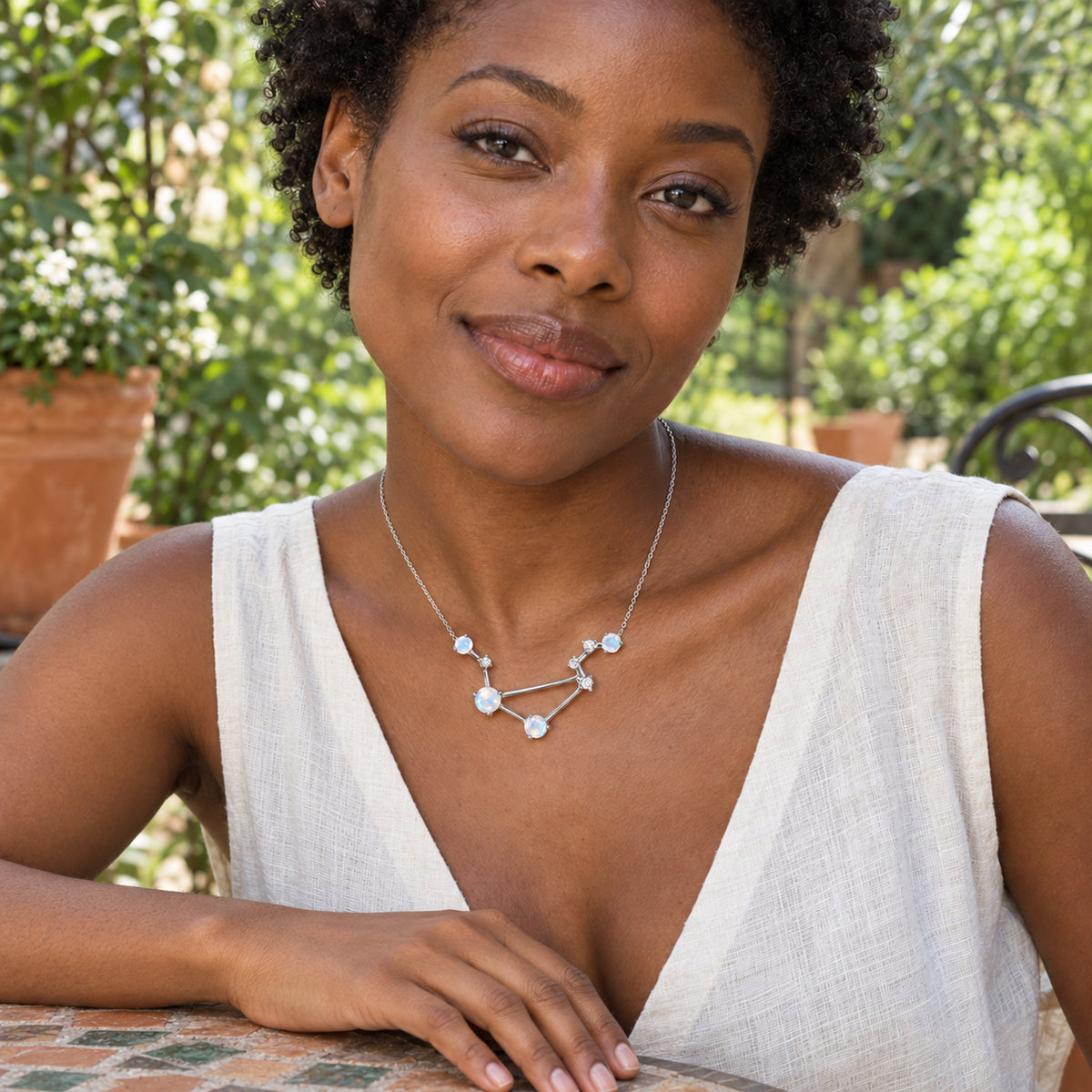 A woman in a white linen top wears a silver moonstone necklace while seated at a mosaic table in a leafy courtyard.