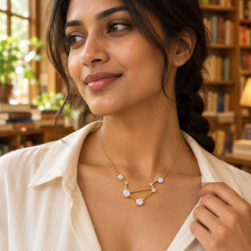 A woman in a cream shirt wears a yellow gold moonstone necklace beside a window and bookshelves.