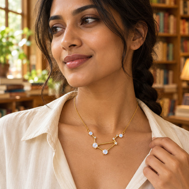 A woman in a cream shirt wears a yellow gold moonstone necklace beside a window and bookshelves.