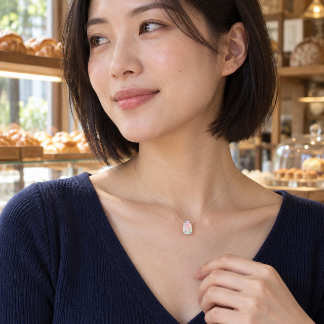 A woman with a dark bob wears a pear-shaped opal in silver prongs on a transparent cord beside a bakery window.
