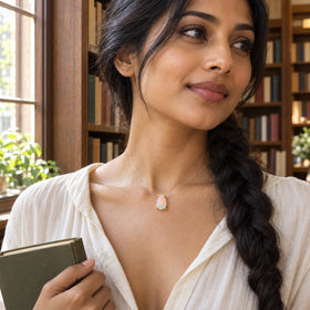 A woman with a long dark braid wears an opal held by yellow gold prongs while holding a green book near wooden shelves.