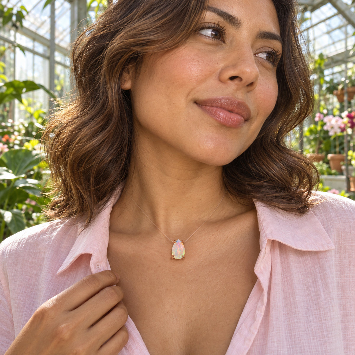 A woman in a pale pink shirt wears a pear-shaped opal with rose gold prongs among sunlit plants in a greenhouse.