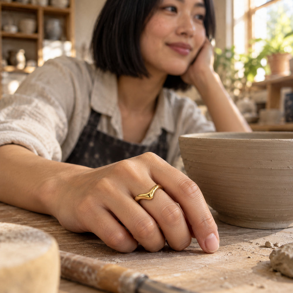 A yellow gold V-shaped ring is worn on a hand resting beside a clay bowl.