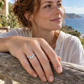 A silver curved band is shown on a freckled hand above a wooden railing with the sea behind it.