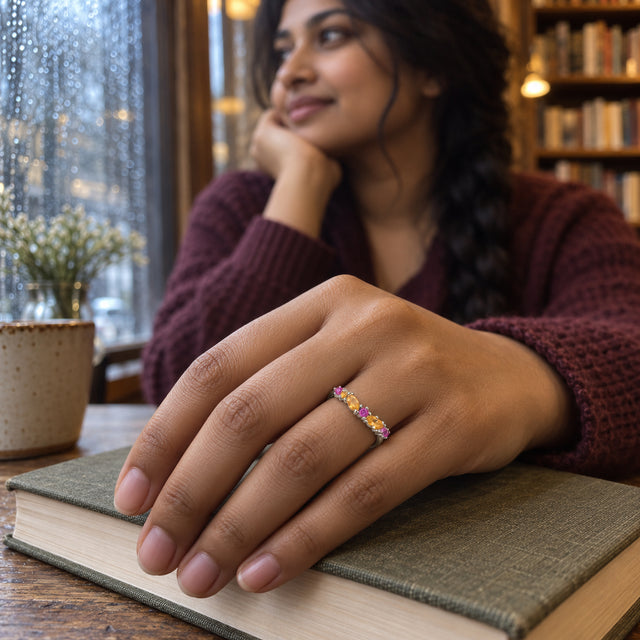 The sterling silver ring with orange and pink gemstones is worn on a hand resting on a cloth-covered book beside a rainy window.