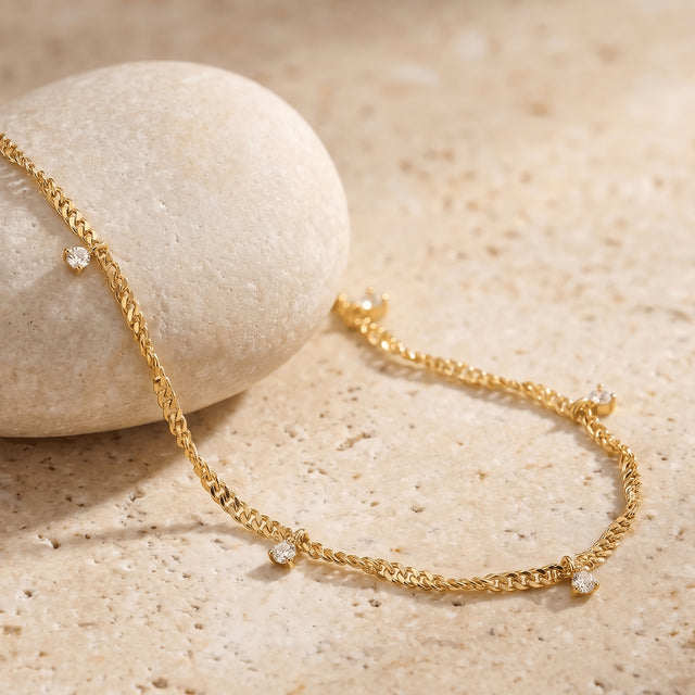 A yellow gold chain and small clear gemstone drops drape across a rounded stone and a cream tabletop.