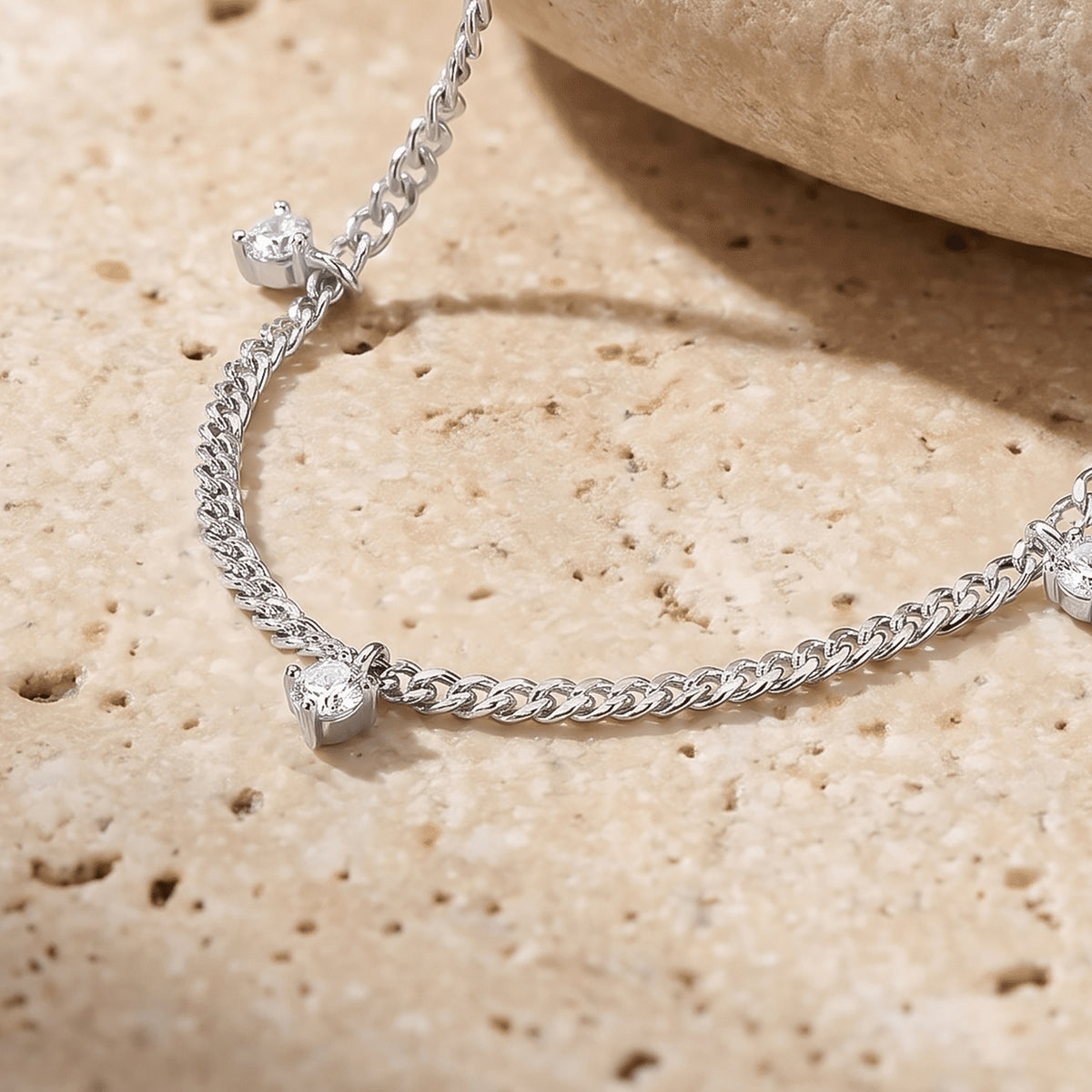 Small clear round gemstone drops hang from a silver chain resting on a textured cream stone surface.