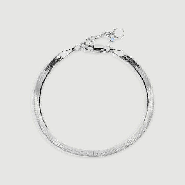 A silver herringbone bracelet forms a rounded loop with its clasp and extension chain visible on a light gray surface.