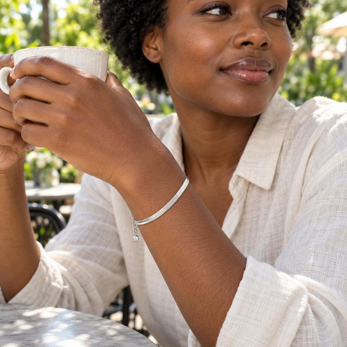 A silver herringbone bracelet wraps around a woman's wrist as she holds a ceramic cup at an outdoor cafe.