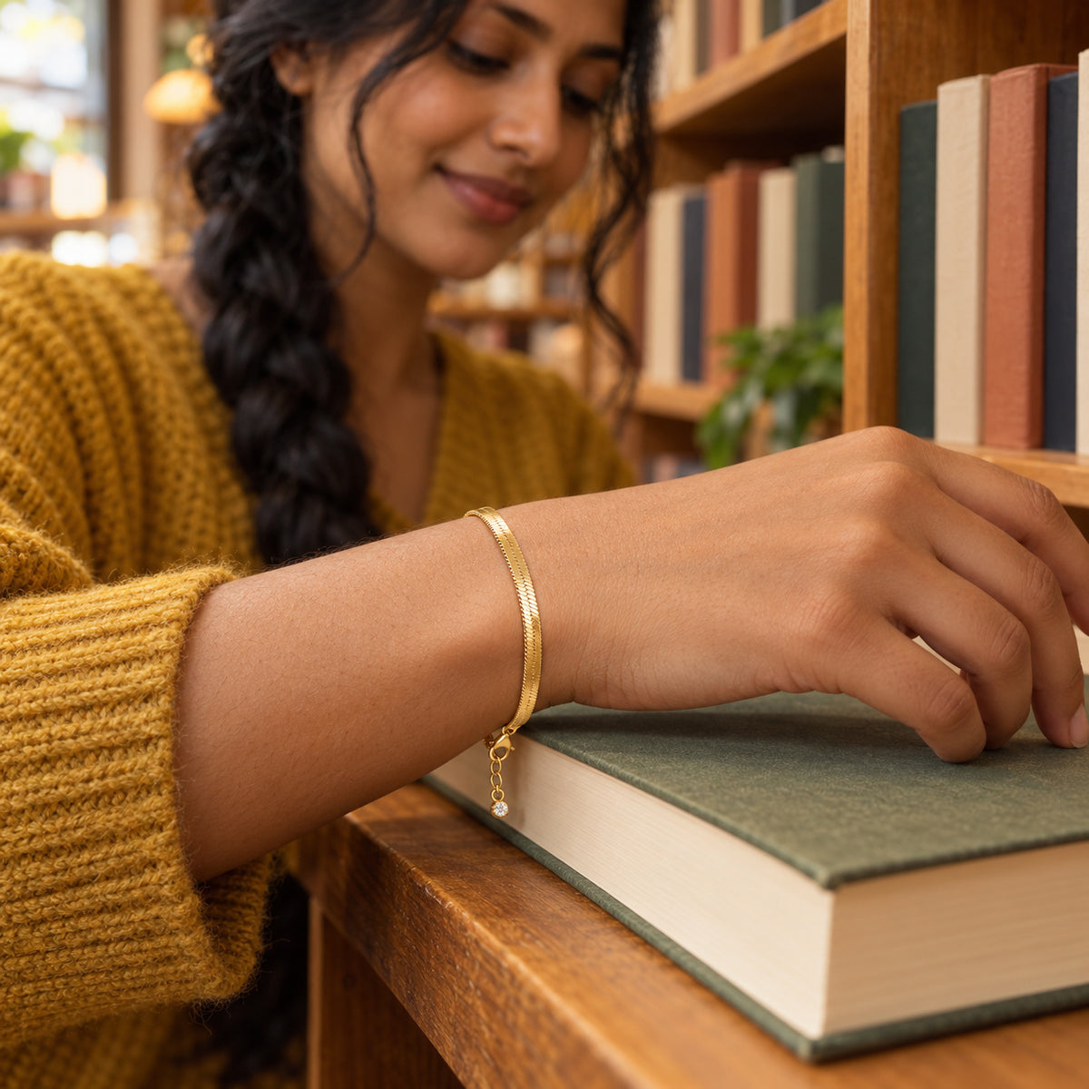 A yellow gold herringbone bracelet is shown on a wrist resting on a green book beside wooden shelves.